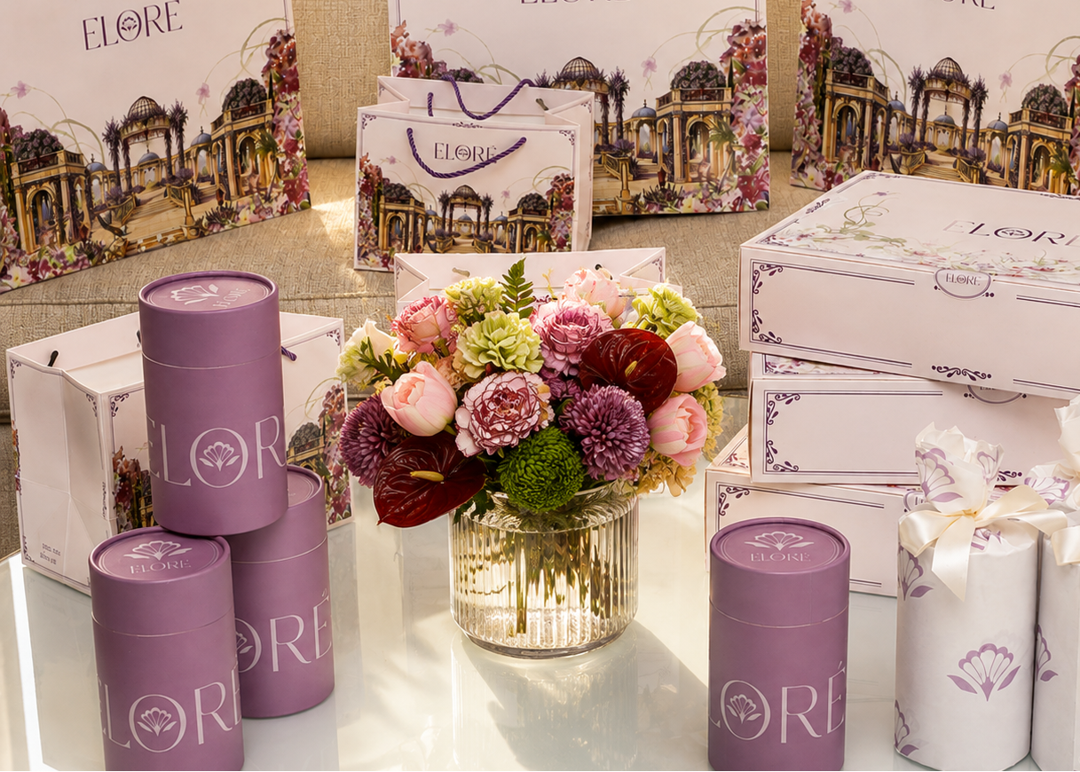 A collection of ELORE skincare and beauty gift items arranged on a glass table in front of a sofa, including purple cylindrical tins, white and lavender gift boxes, and a central floral arrangement.