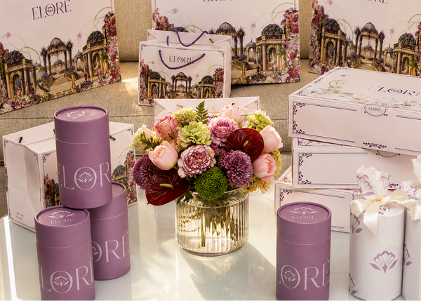 A collection of ELORE skincare and beauty gift items arranged on a glass table in front of a sofa, including purple cylindrical tins, white and lavender gift boxes, and a central floral arrangement.