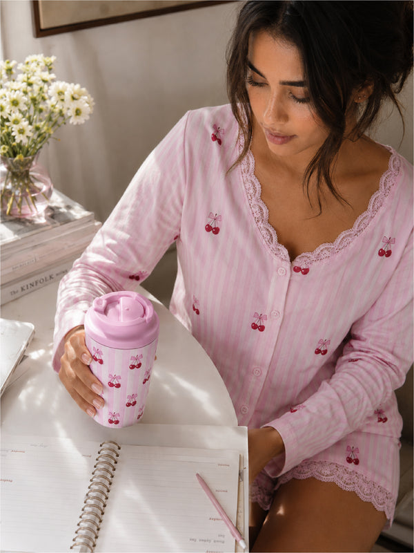 Woman in pink pajama set holding a pink travel mug with cherry design, sitting at a desk.