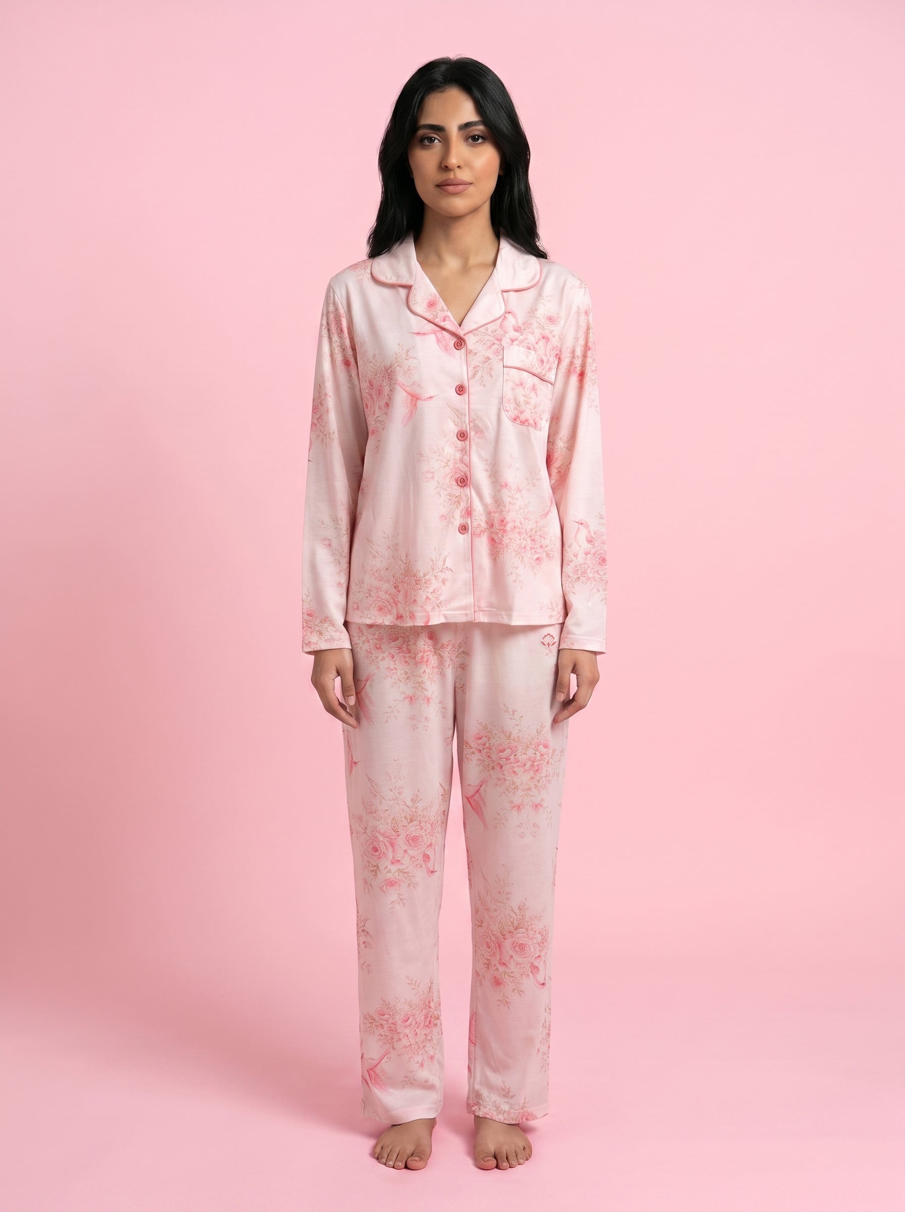 A woman modeling a light pink long-sleeve pajama set with a soft pink floral print, standing against a solid pink studio background.