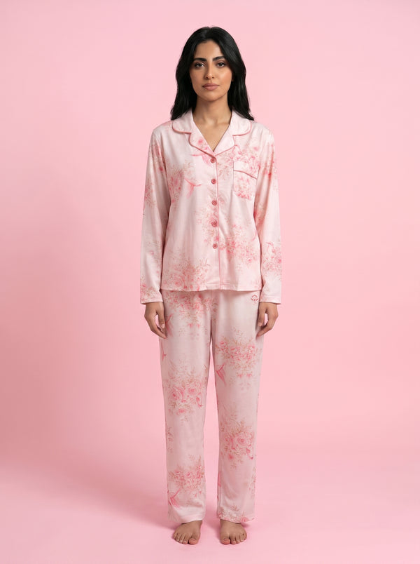 A woman modeling a light pink long-sleeve pajama set with a soft pink floral print, standing against a solid pink studio background.